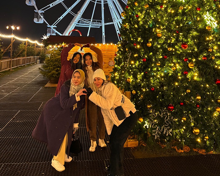 Sharu and friends enjoy a festive evening at a Christmas market, surrounded by decorations and a large Ferris wheel.