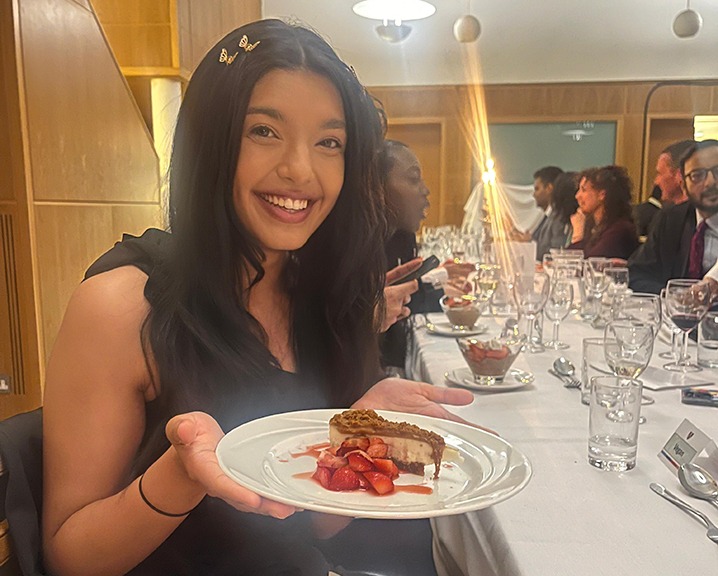 Sharu Iyer holds a dessert at a formal dinner, smiling amidst elegantly set tables and fellow attendees.