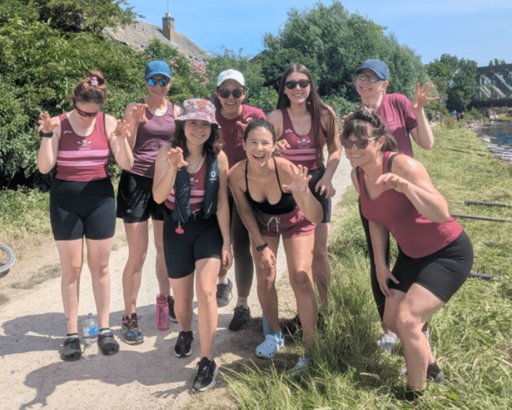 Peiju Li and her rowing team pose cheerfully by the river in matching maroon attire on a sunny day.